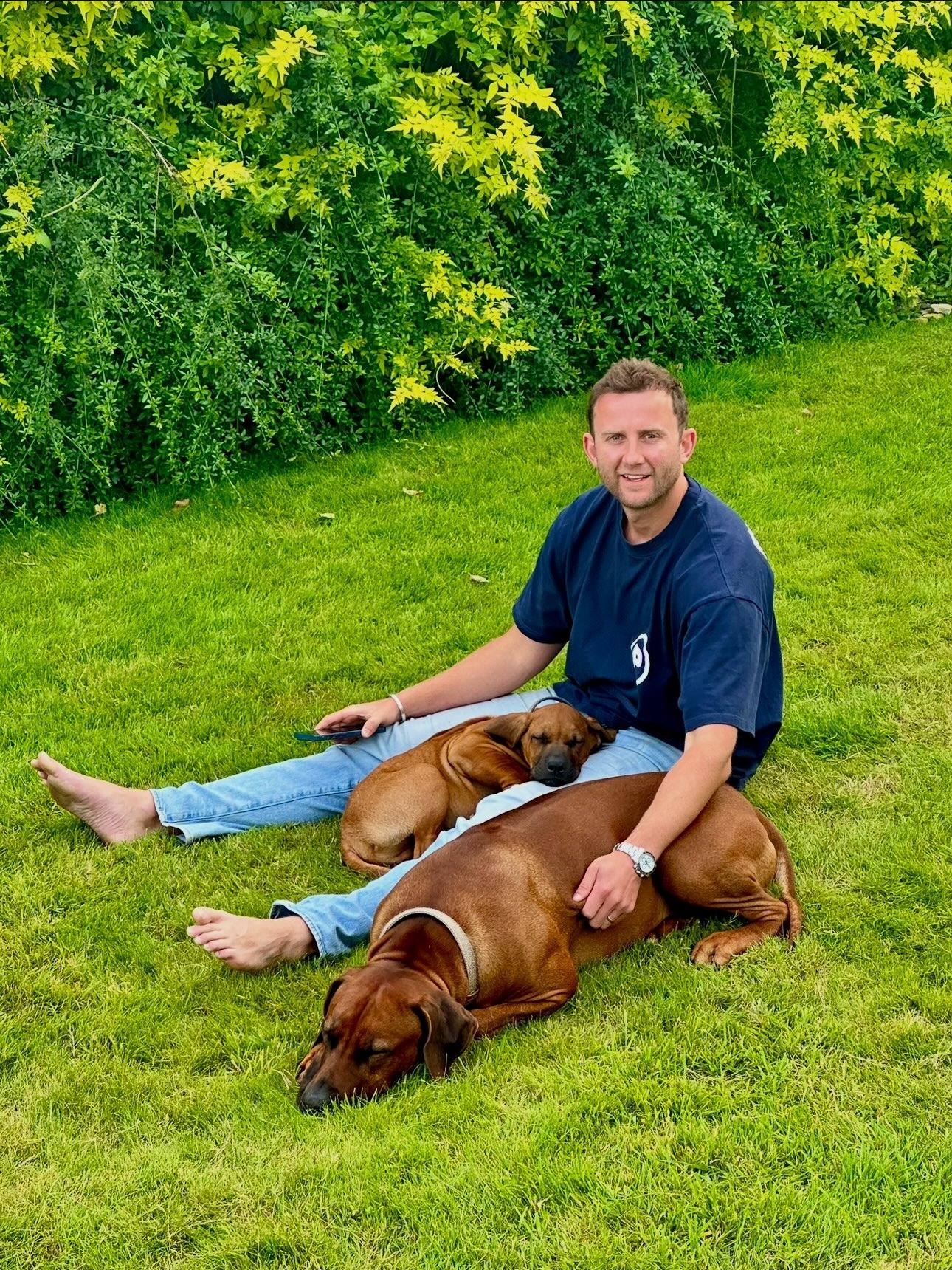 Hugo sitting barefoot on the lawn with two Rhodesian ridgebacks asleep across his legs.